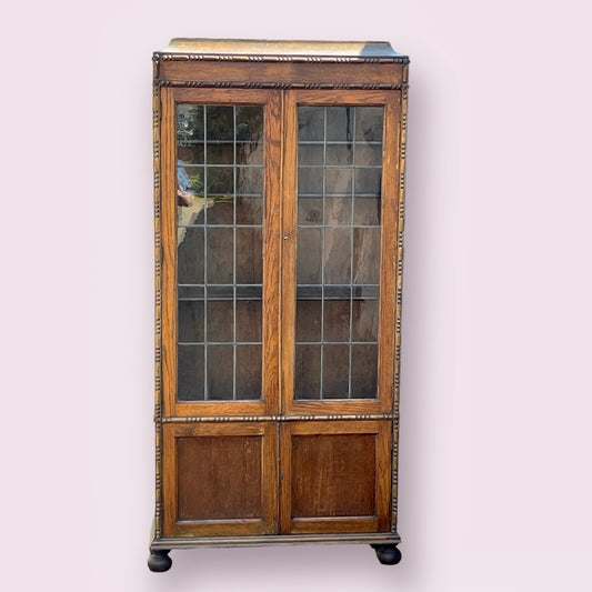 Tall, slim, vintage, wood-crafted, glazed book/china display cabinet features two leaded glass doors with three adjustable wooden shelves. With lower storage cabinet beneath the doors.